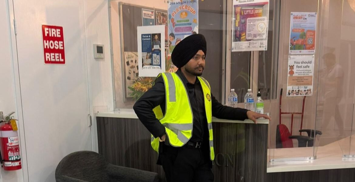 Security guard in high-visibility vest at gatehouse with fire hose reel sign, overseeing entrance and ensuring safety at a busy site.