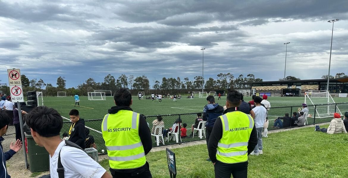 Security personnel from AS Security monitoring a soccer event, wearing high-visibility vests, with spectators and a soccer field in the background.