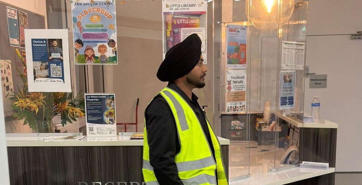 Security guard in a high-visibility vest at a reception area, surrounded by informational posters about community programs and safety, representing AS Security's mobile patrol services.