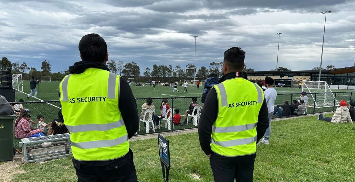 Security personnel from AS Security monitoring a public soccer event, wearing high-visibility vests, with spectators and a soccer field in the background.