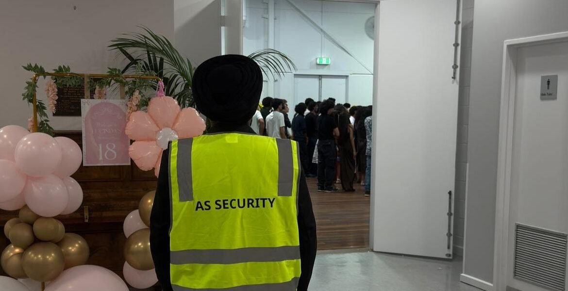 Security guard in a high-visibility vest labeled "AS SECURITY" overseeing a public event with decorative balloons and guests in the background.