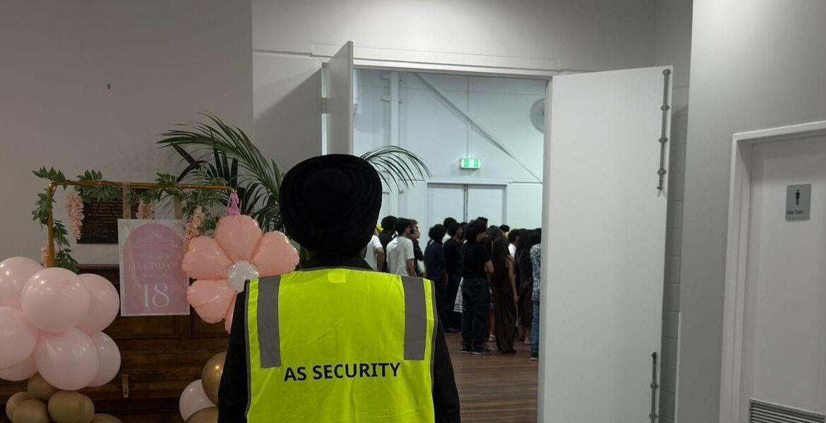 Security guard in yellow vest with "AS SECURITY" text overseeing a public event with attendees in the background, decorated with balloons and floral arrangements.